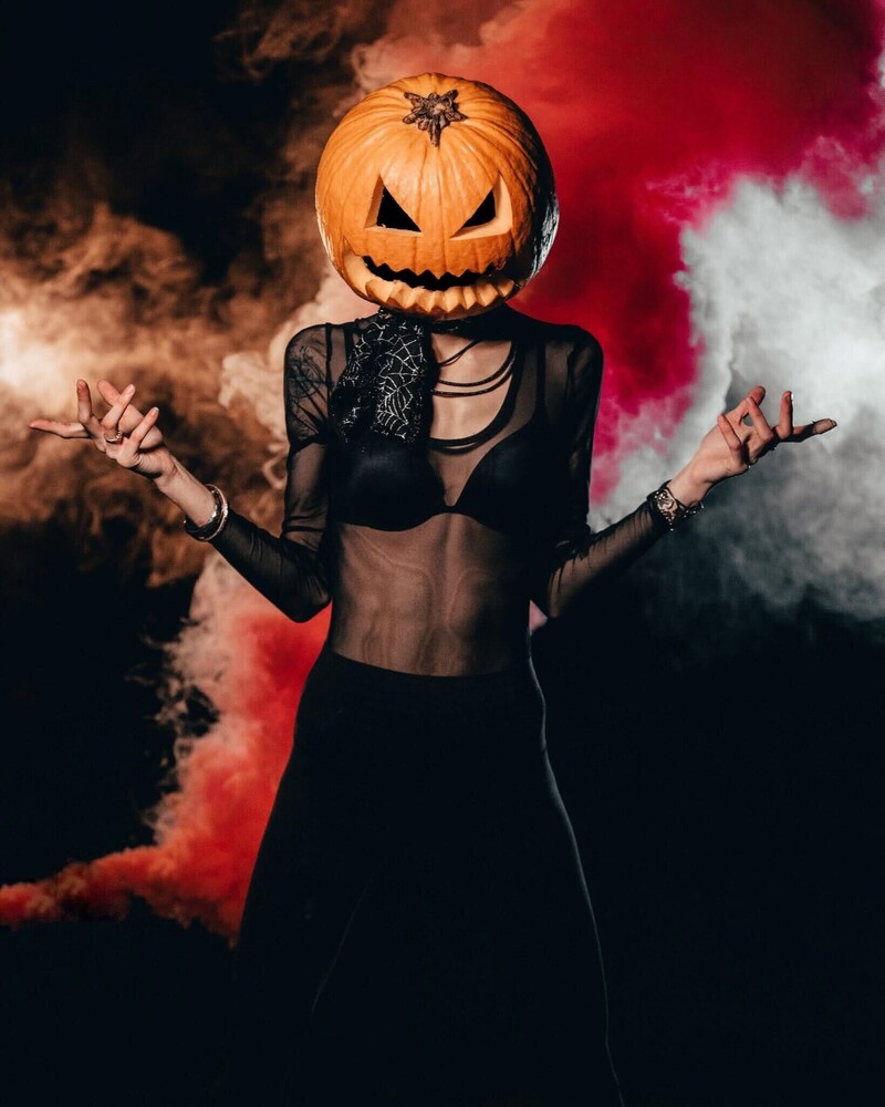 Person wearing jack-o'-lantern pumpkin head and black crop top with arms raised in dramatic pose