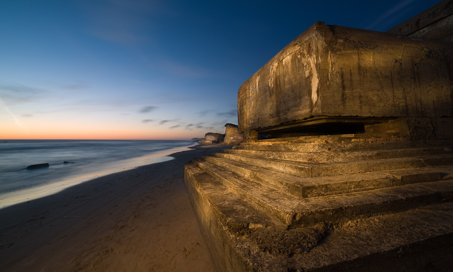 Weathered concrete coastal bunker with steps on a sandy beach at dusk, with smooth long-exposure surf to the left.
