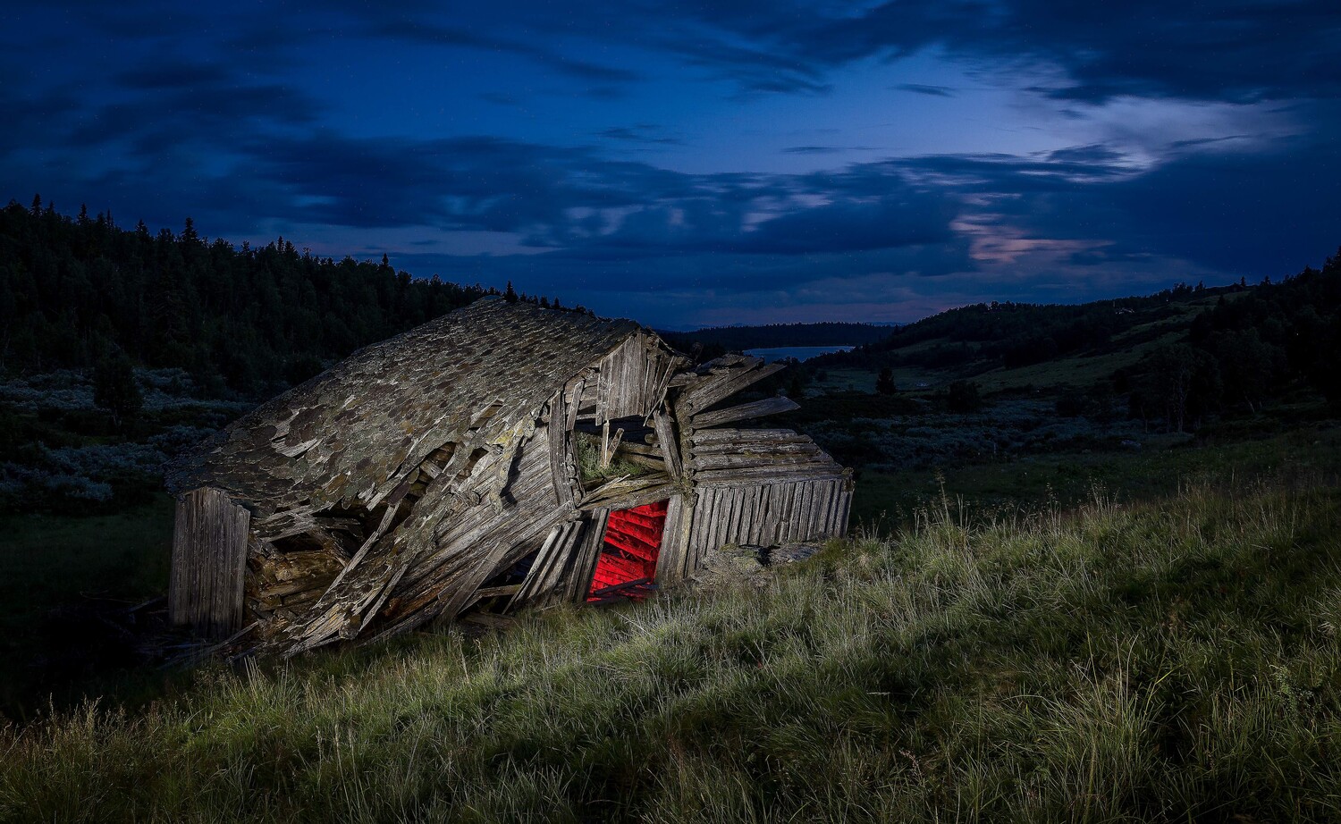 Dilapidated wooden barn leaning on a grassy hillside under a blue dusk sky, its single doorway glowing red.