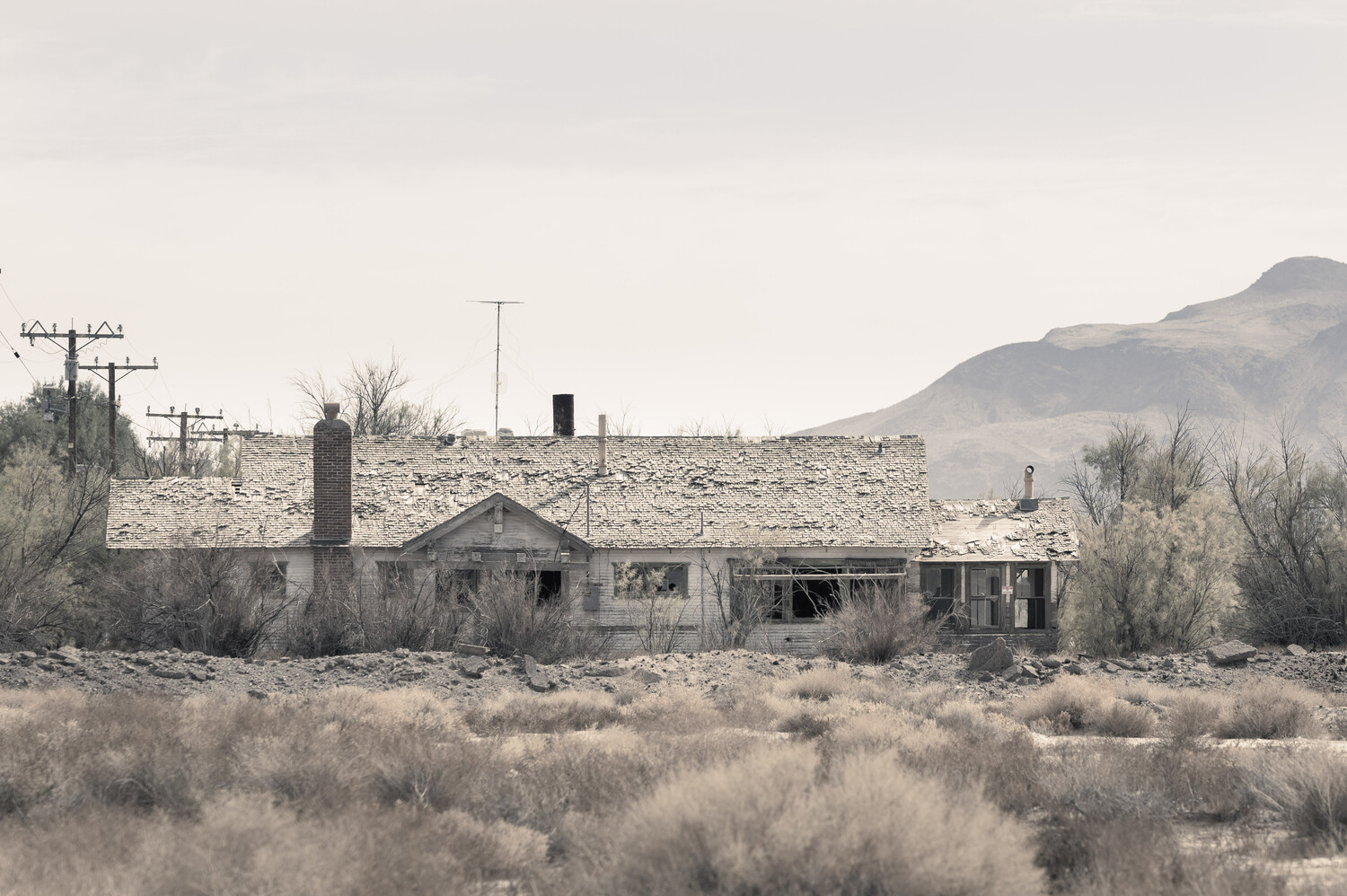Weathered abandoned single-story building with a collapsed roof and brick chimney in a desert landscape, with a mountain ridge behind it.