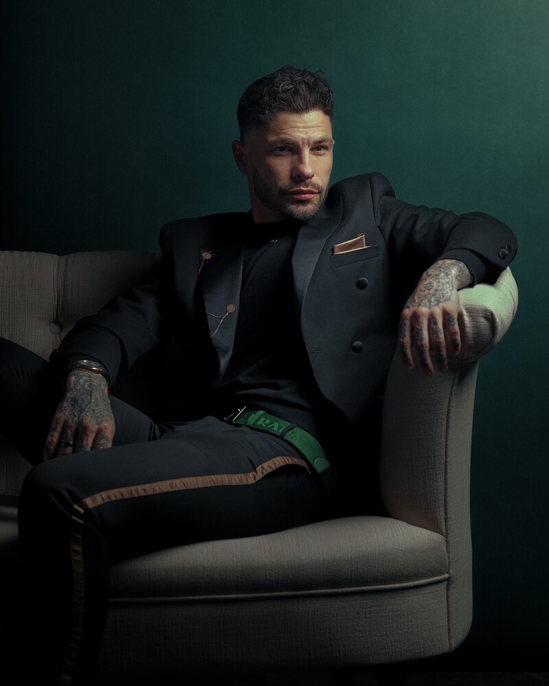 Man in dark blazer seated on gray armchair against teal background