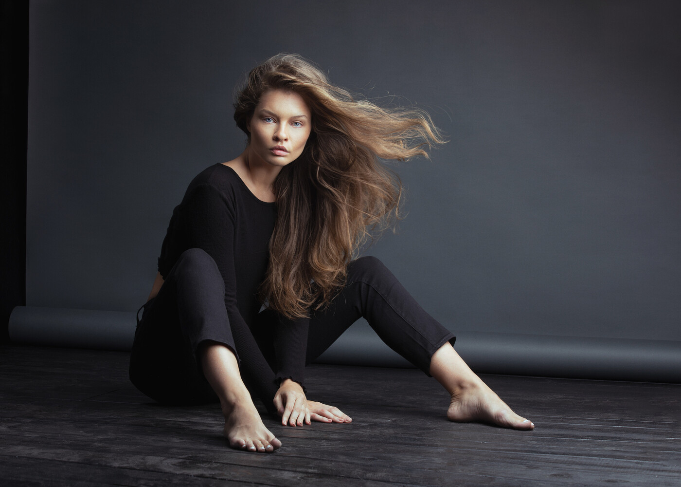 Woman in black clothing seated on dark floor with long brown hair flowing