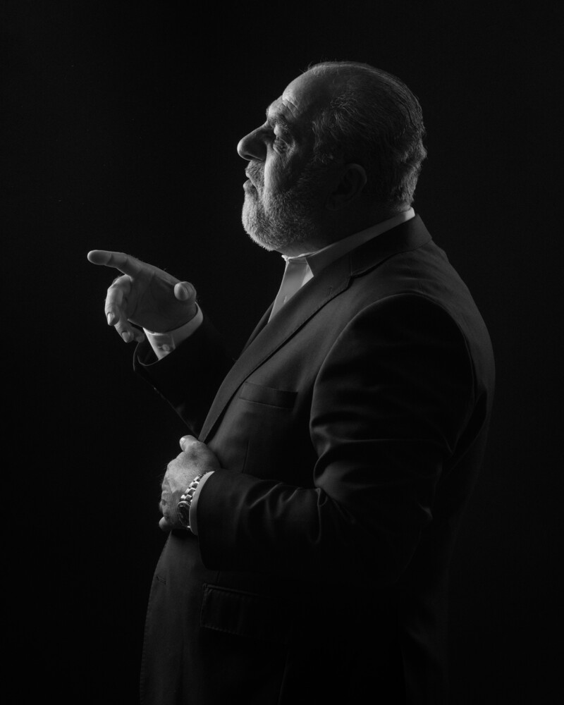 Bald man in dark suit gesturing while speaking against black background
