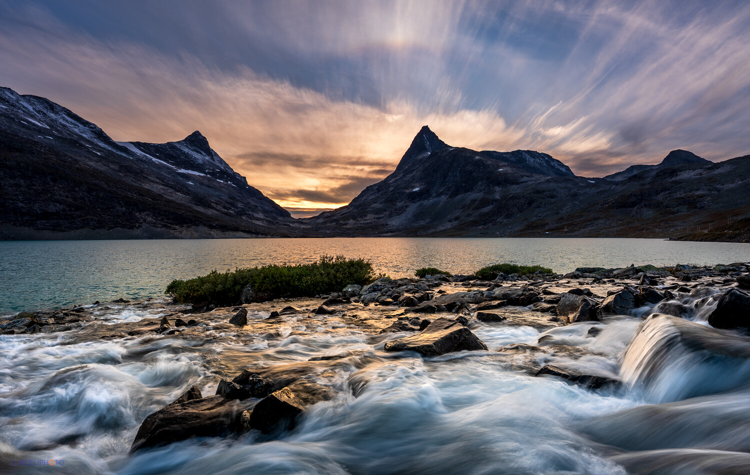 Snow-capped mountains frame a glacial lake with turbulent water and rocky shores at golden hour