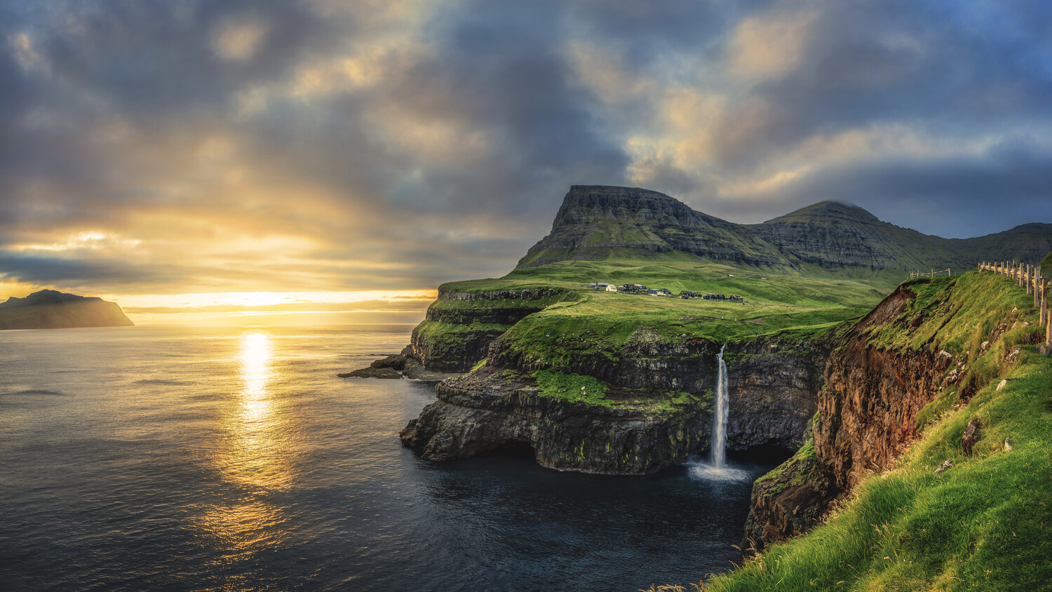 Waterfall cascading from green cliffs onto rocky coastline at golden hour sunset