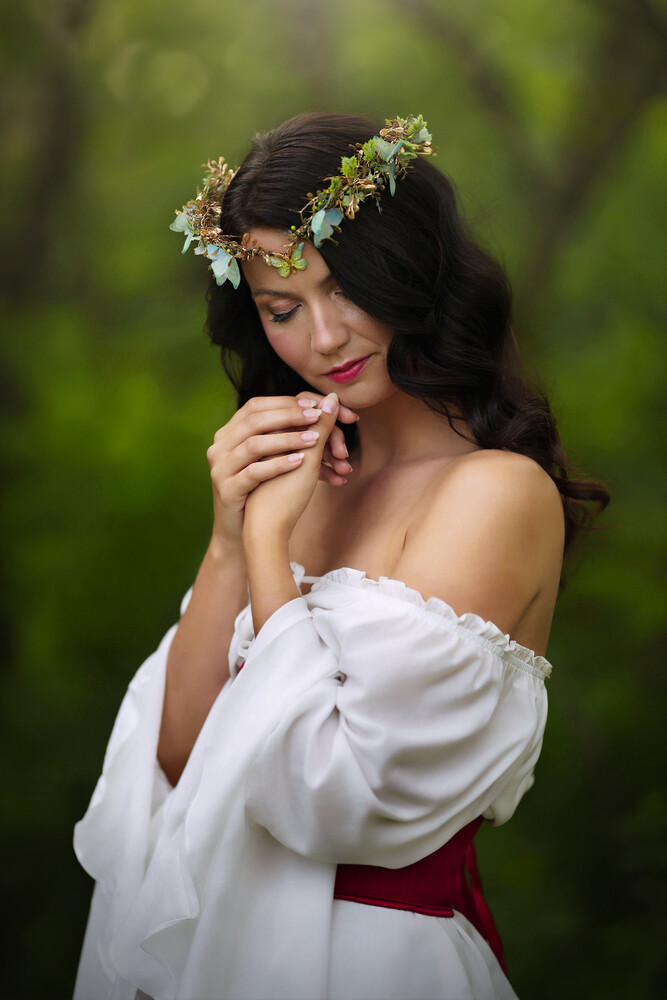 Woman in white off-shoulder blouse wearing floral crown, posed against soft green bokeh background