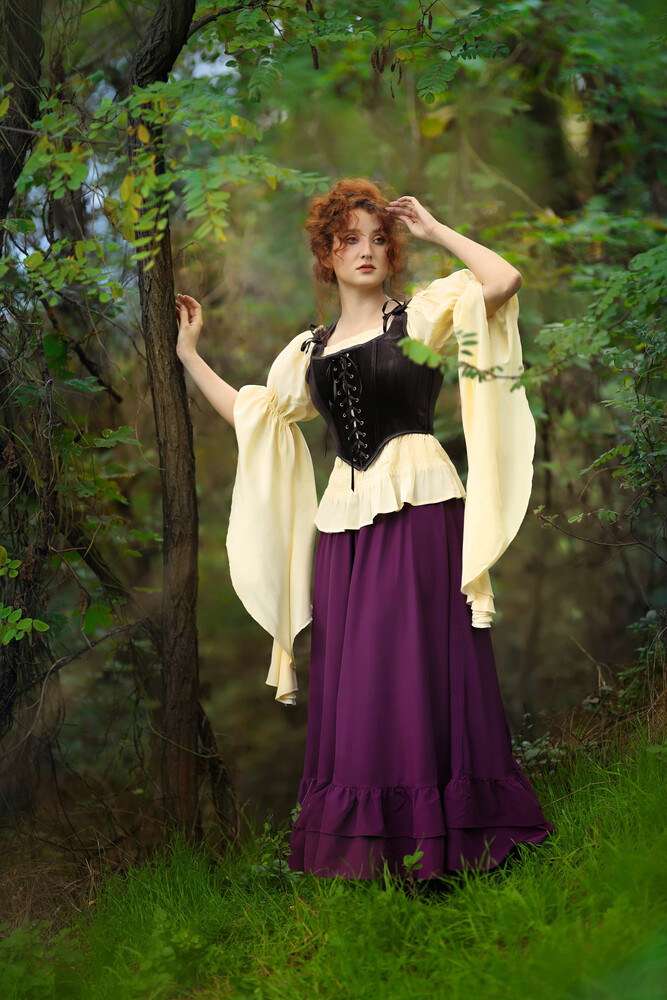 Woman in Renaissance-style costume standing among green foliage, adjusting her curled hair