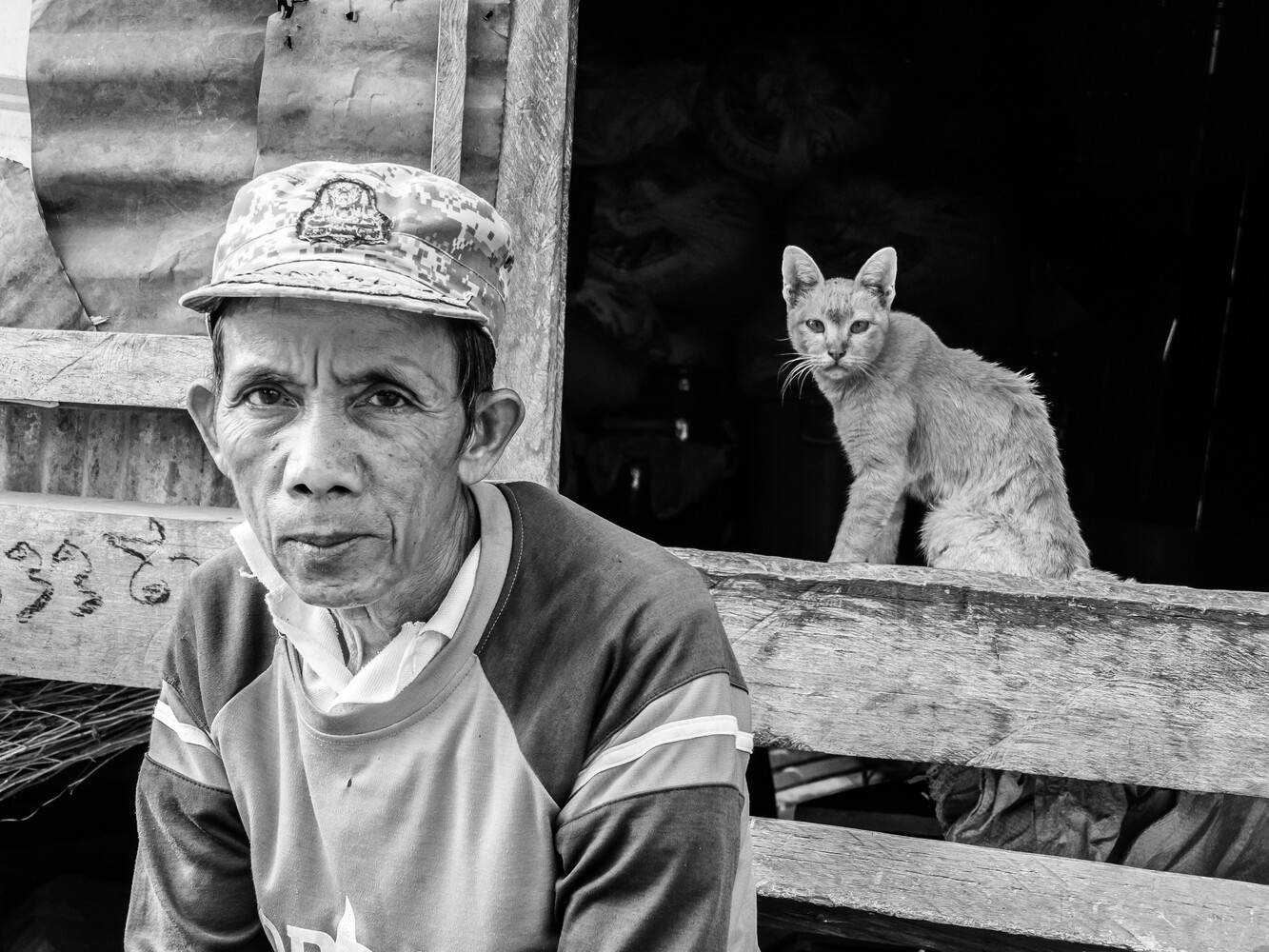 Man in cap and casual shirt poses beside wooden railing with alert cat perched behind him