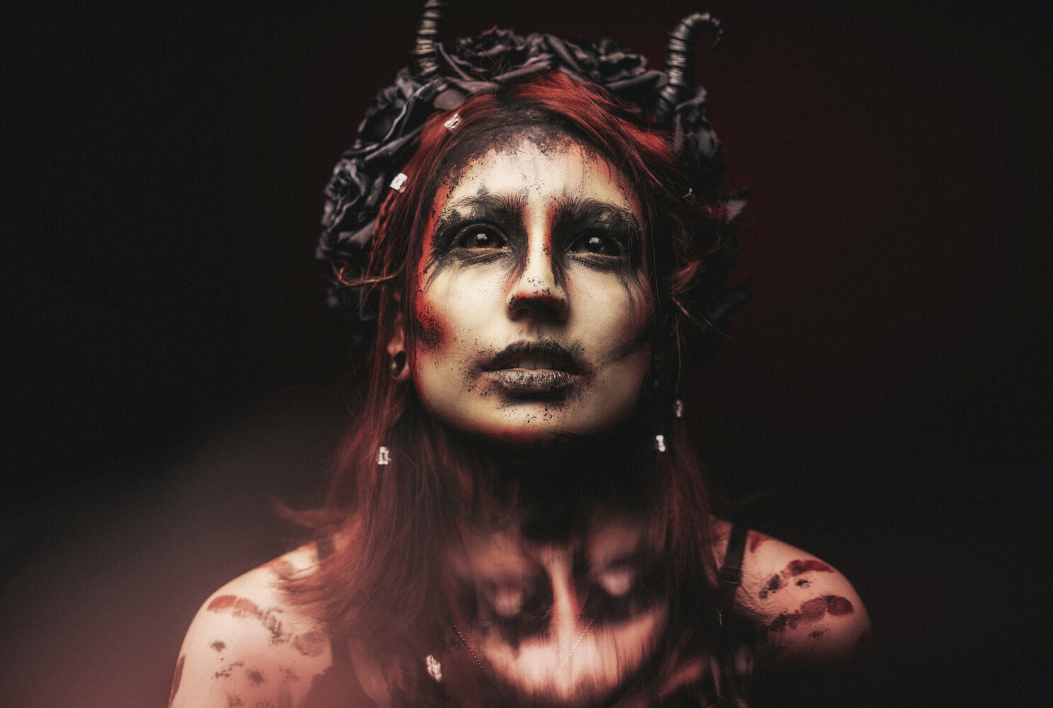 Woman with pale face paint, dark eye makeup, and curved horns wearing a black floral crown against a dark red background.