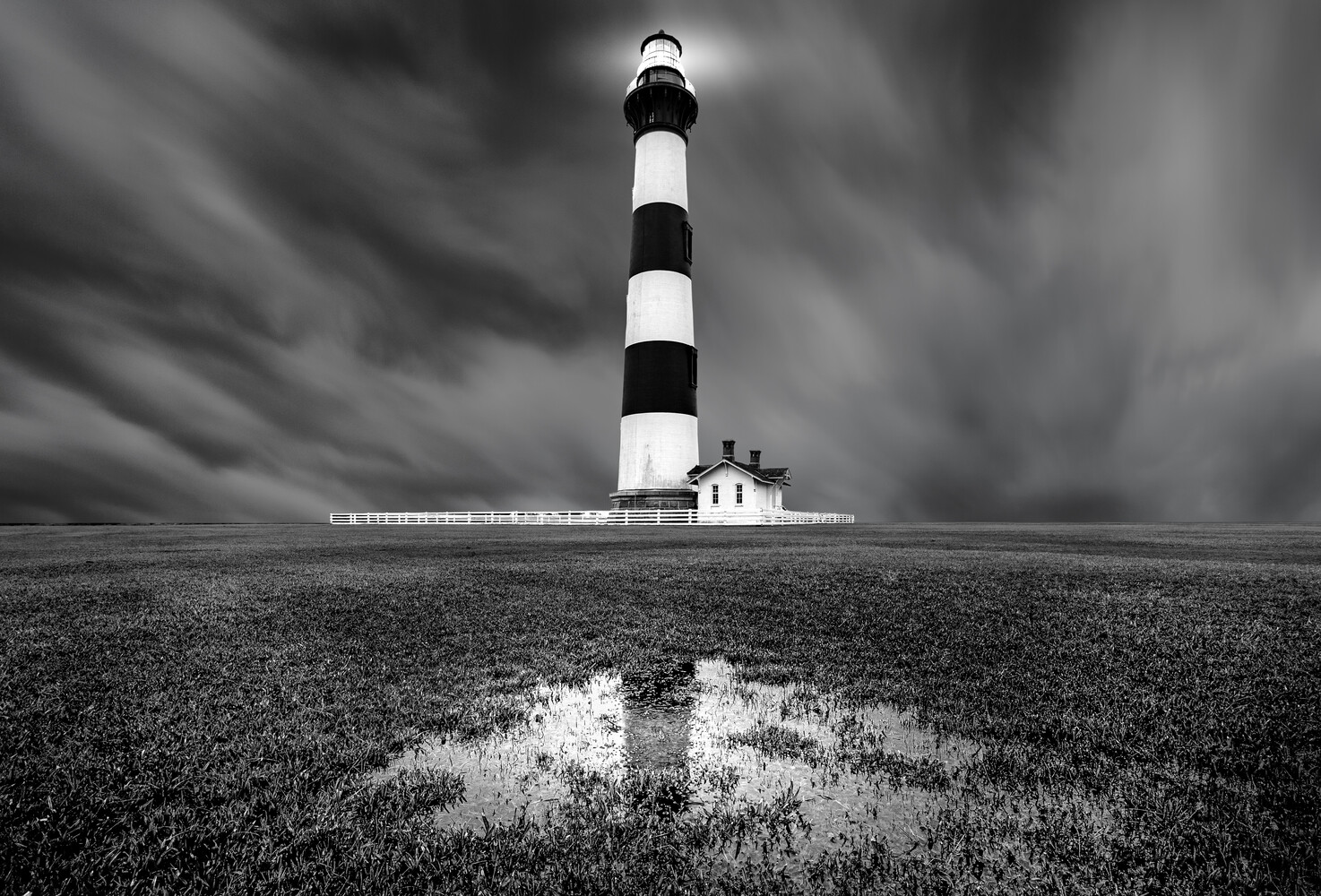 Striped lighthouse standing alone in a vast, flat landscape with a puddle reflection and dramatic storm clouds