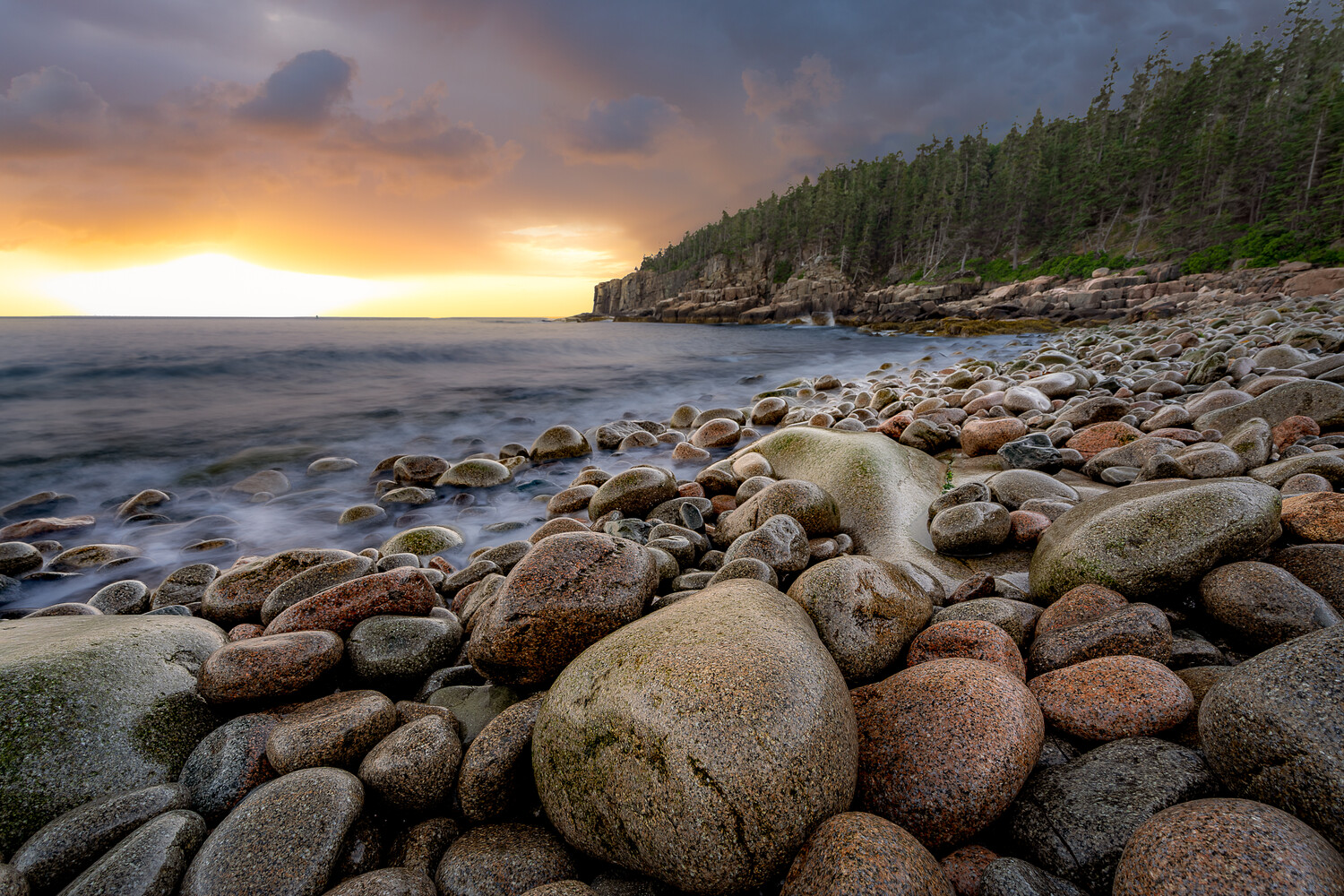 Rocky shoreline at sunset with forested cliffs and golden sky reflecting on calm water