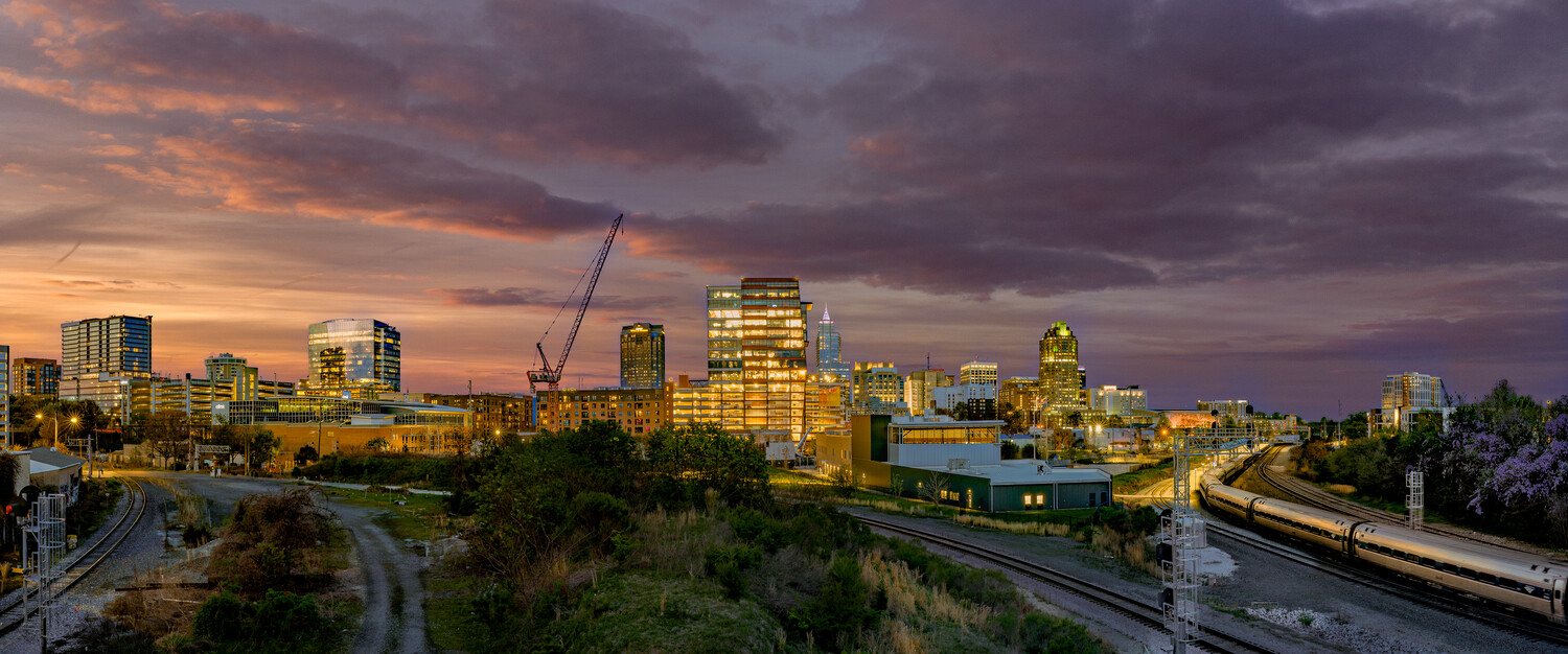 City skyline at dusk with illuminated buildings, railroad tracks, and dramatic purple-orange sky