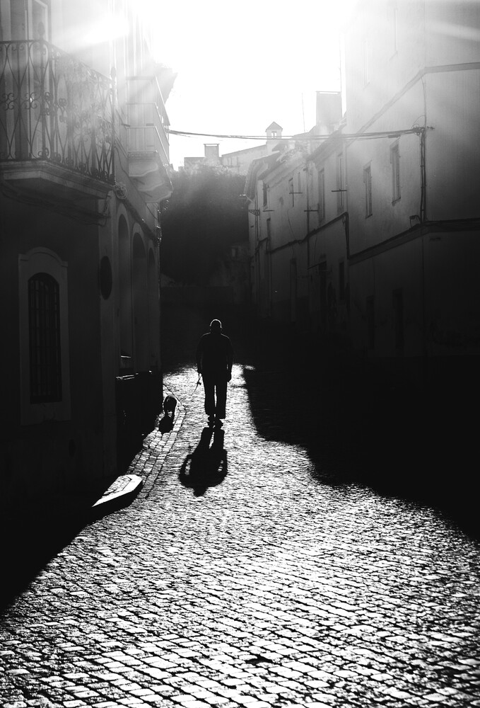 Solitary figure in dark coat walking down narrow cobblestone alley between tall buildings