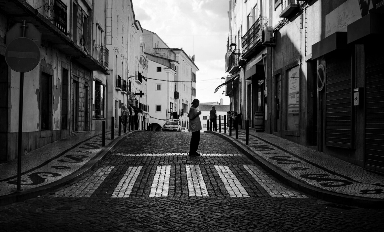 Black and white street scene of a person standing in the middle of a cobblestone crosswalk in a narrow old town street.