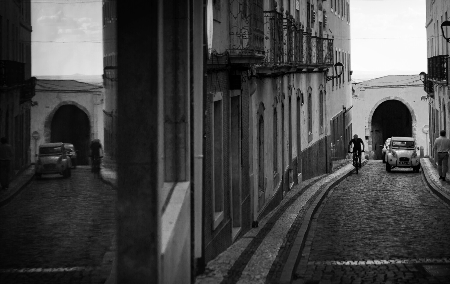 Black and white view of a cyclist riding down a narrow cobblestone street toward an arched gateway in an old town.