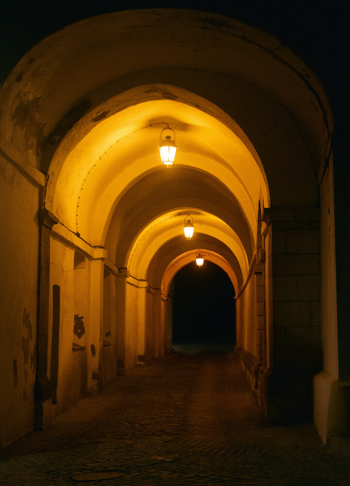 Long stone arcade tunnel lit by warm hanging lanterns, receding toward a dark opening at the end of the passage.