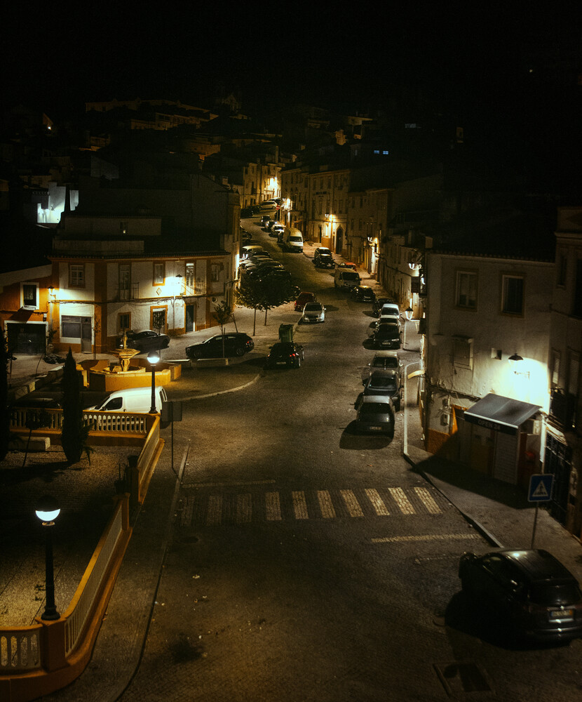 Night view down a narrow street lined with parked cars, illuminated by warm streetlamps and building lights in a small town