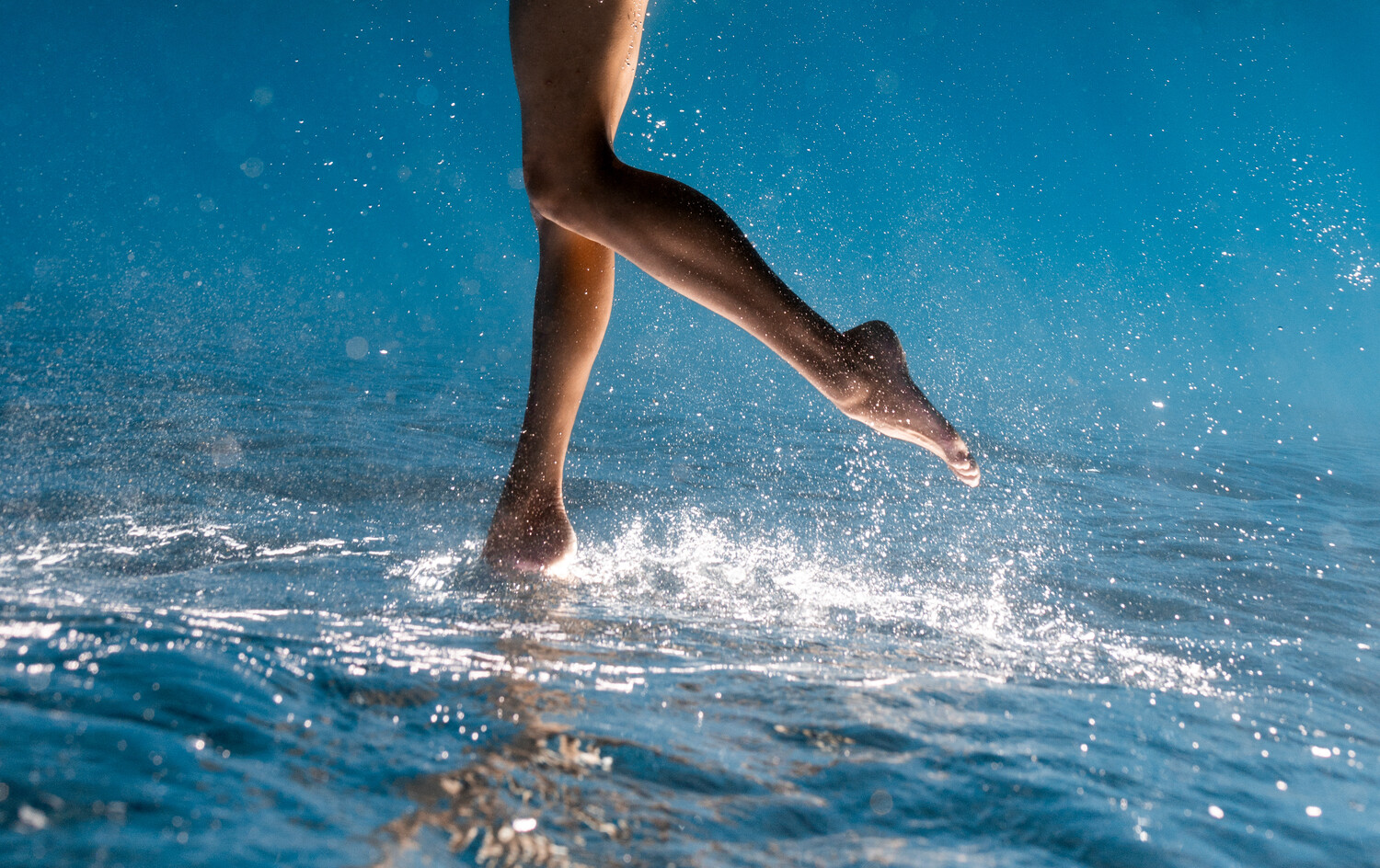 Runner's legs splashing through shallow ocean water in bright sunlight
