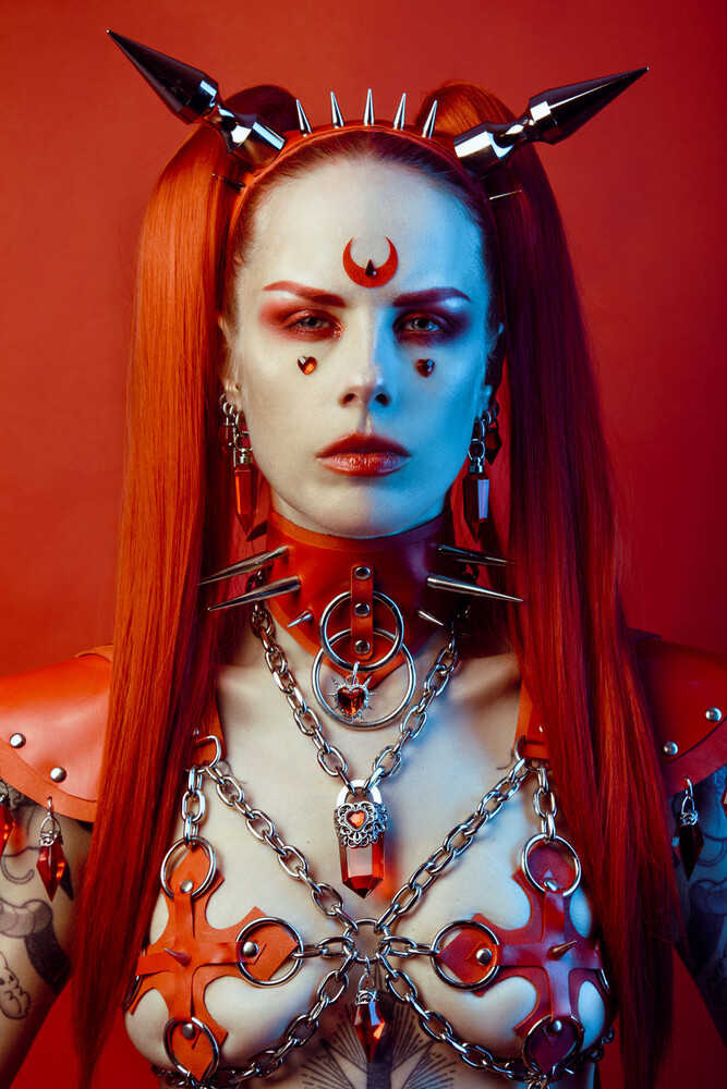 Woman with long red hair in a spiked metal headpiece and chain harness, facing the camera against a red backdrop.
