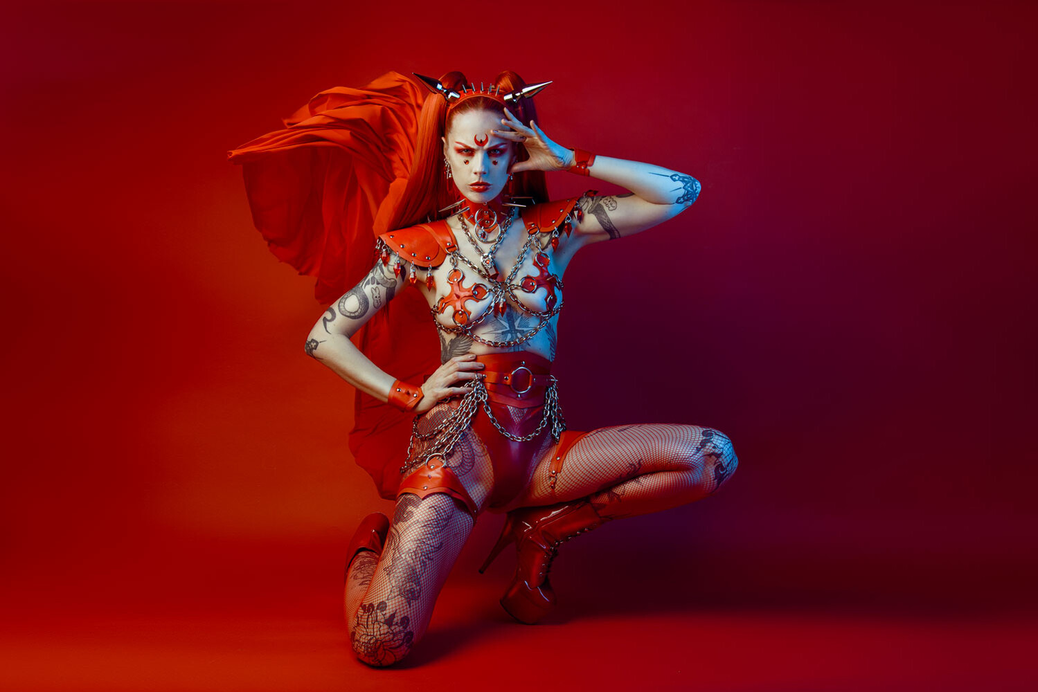 Woman in red fantasy body paint and chain harness kneeling against a red backdrop with flowing red fabric behind her