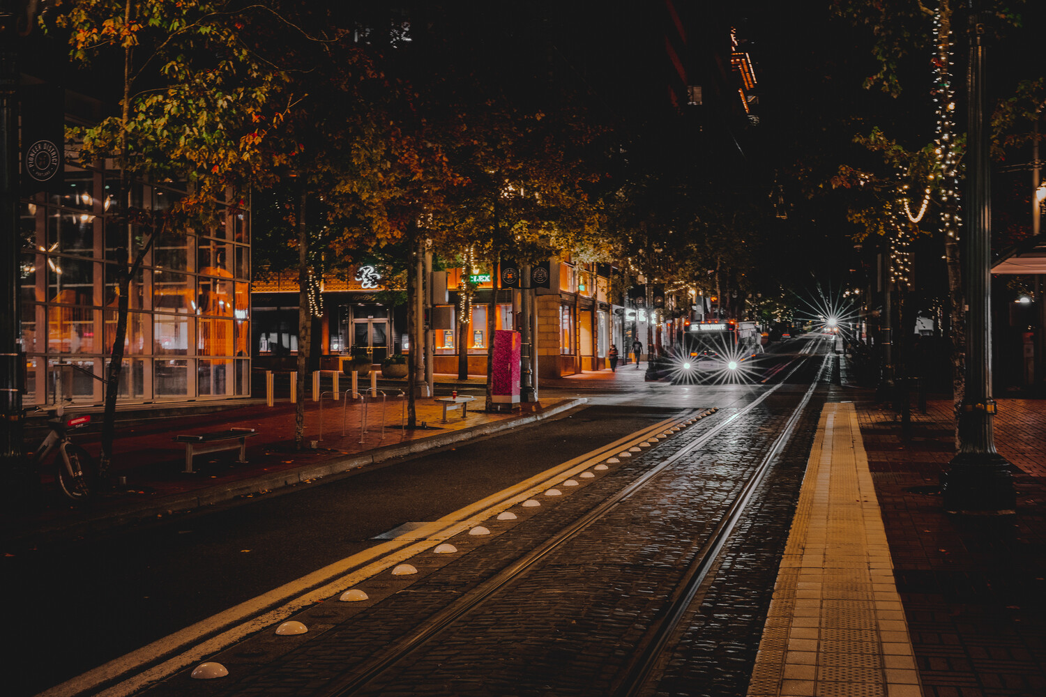 Nighttime urban street with tram tracks, warm storefront lighting, and trees lining wet pavement