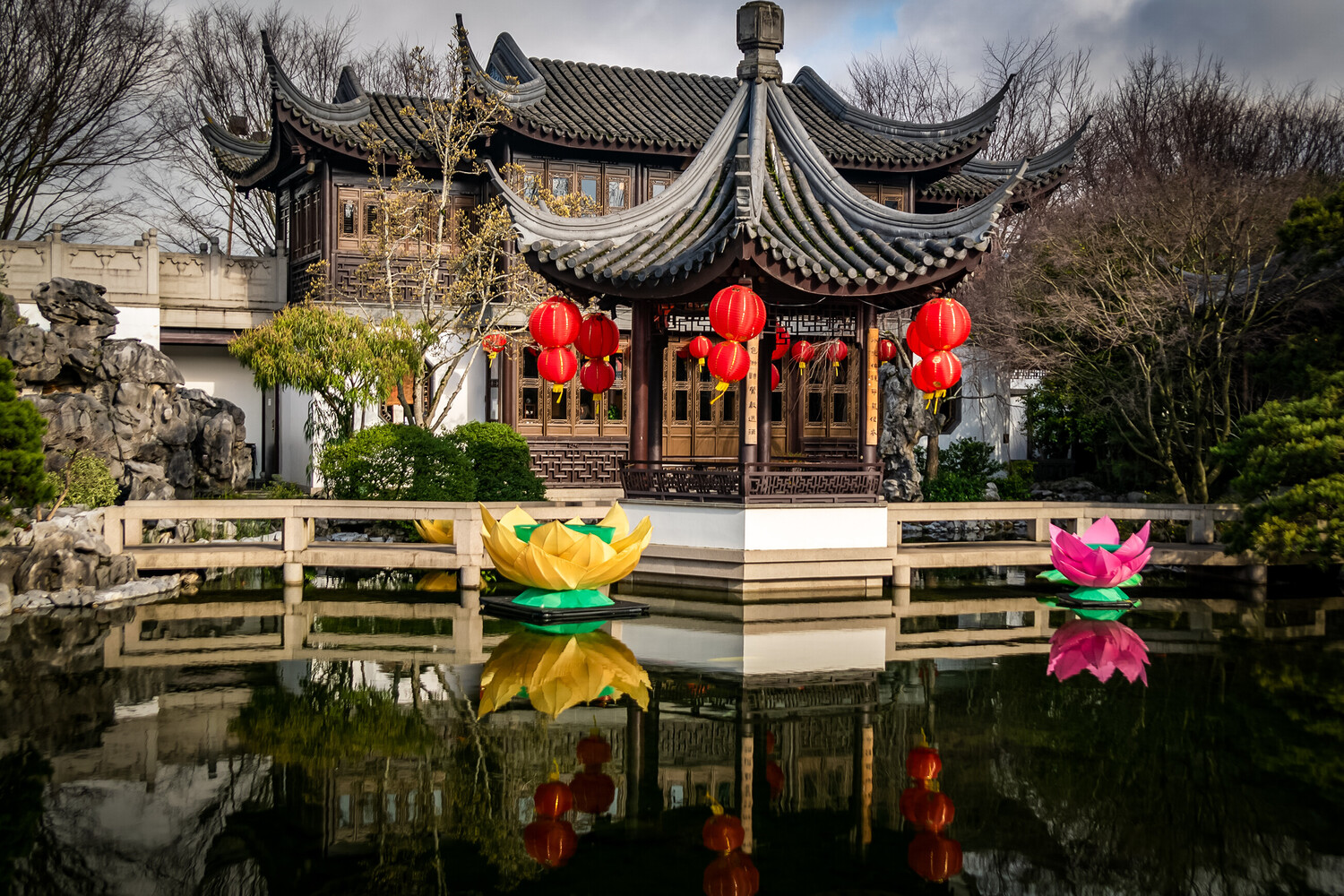 Traditional Chinese pavilion with upturned eaves reflected in still water, flanked by colorful floating lotus flower decorations