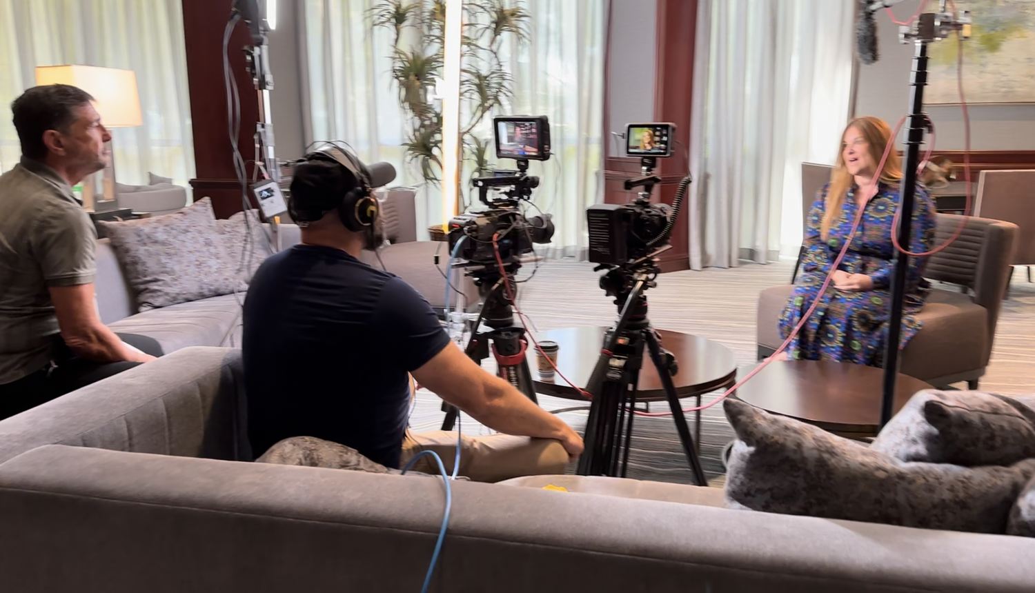 Behind-the-scenes view of an interview shoot, with a woman in a blue patterned dress seated across from cameras on tripods in a hotel-style room.