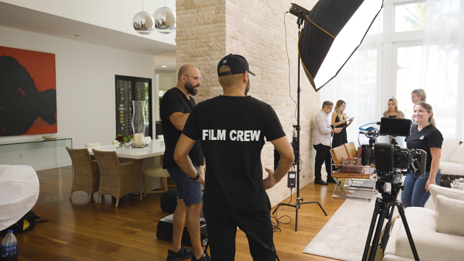 Film crew in black shirts setting up a shoot in a bright modern home, with a softbox light and camera on a tripod in the foreground.