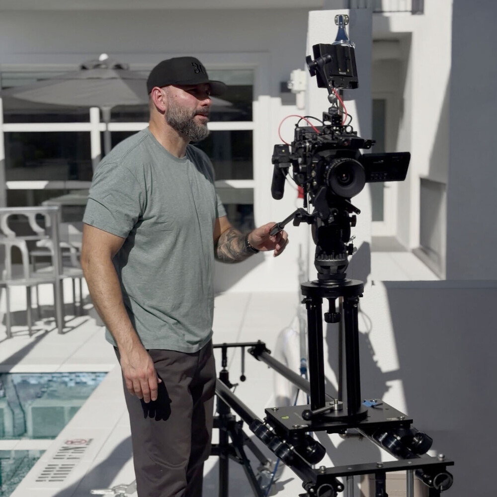 Bearded man in a black cap and grey t-shirt operating a cinema camera on a tripod rig on a sunny white rooftop terrace