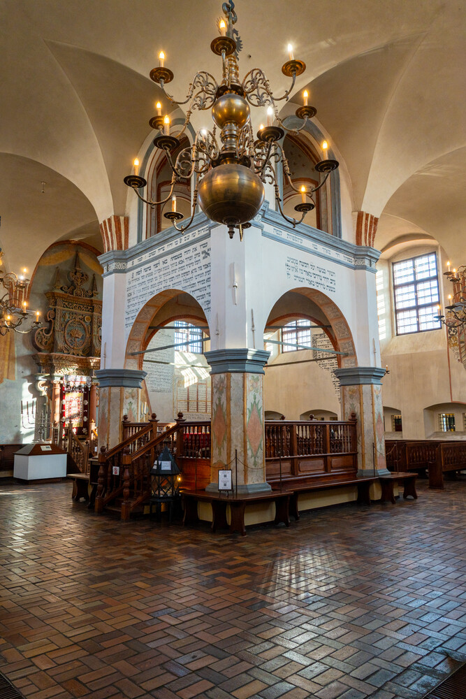 Ornate brass chandelier hanging above a central raised wooden platform with arched stone supports in a vaulted, historic interior