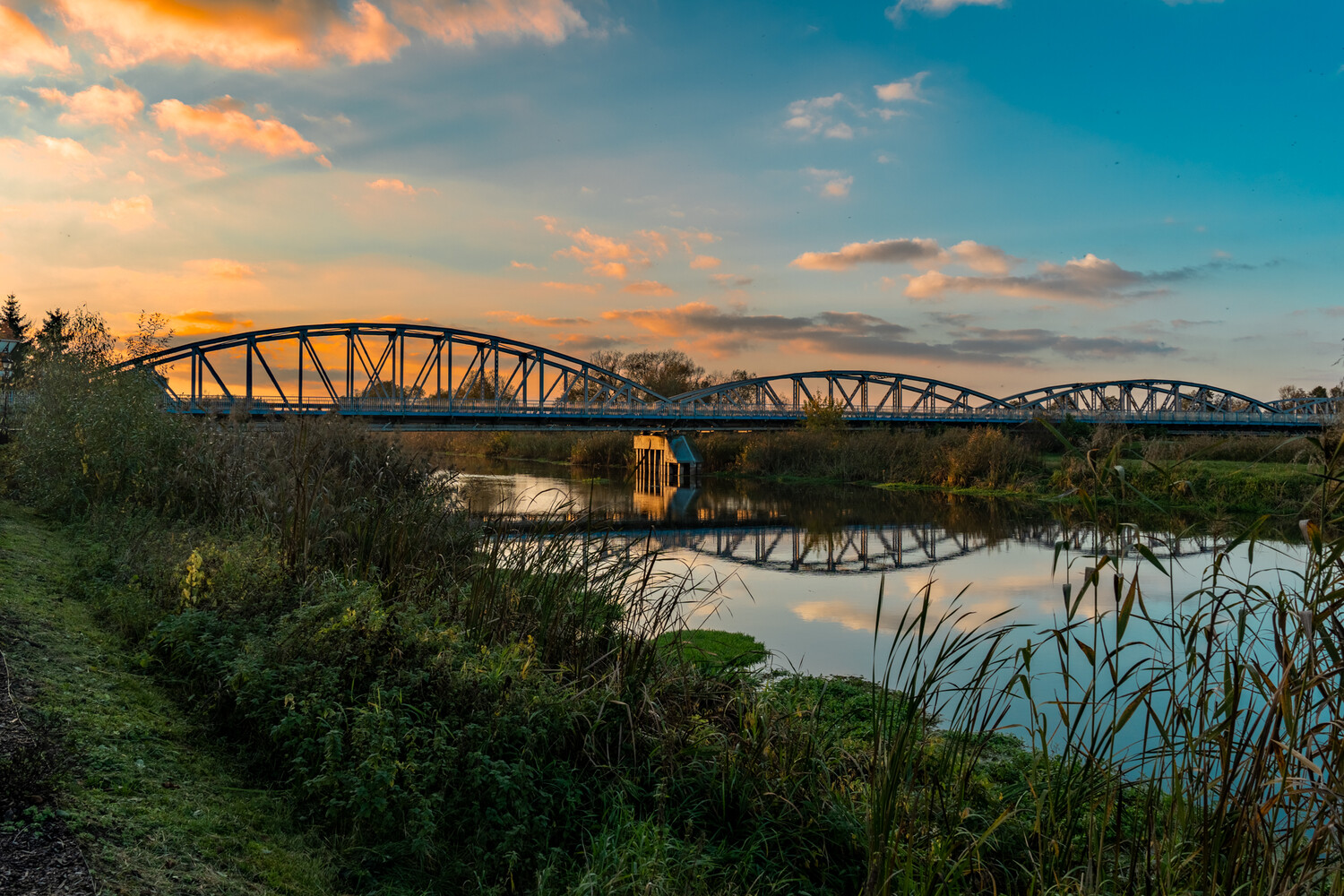 Steel truss bridge arching over a calm river at sunset, its reflection mirrored in the still water beneath golden and blue skies.