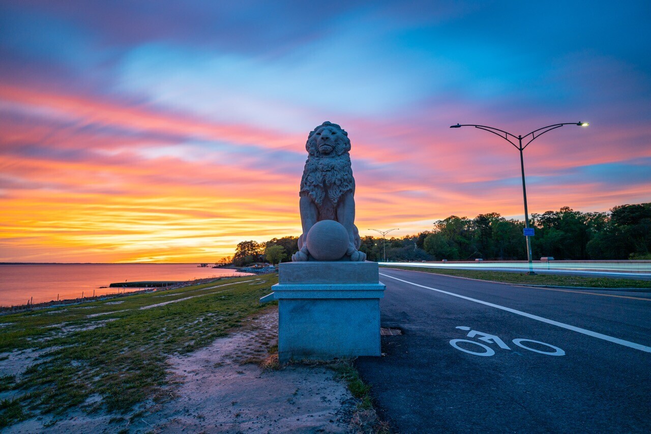 Stone lion statue on a white pedestal beside a coastal road at sunset