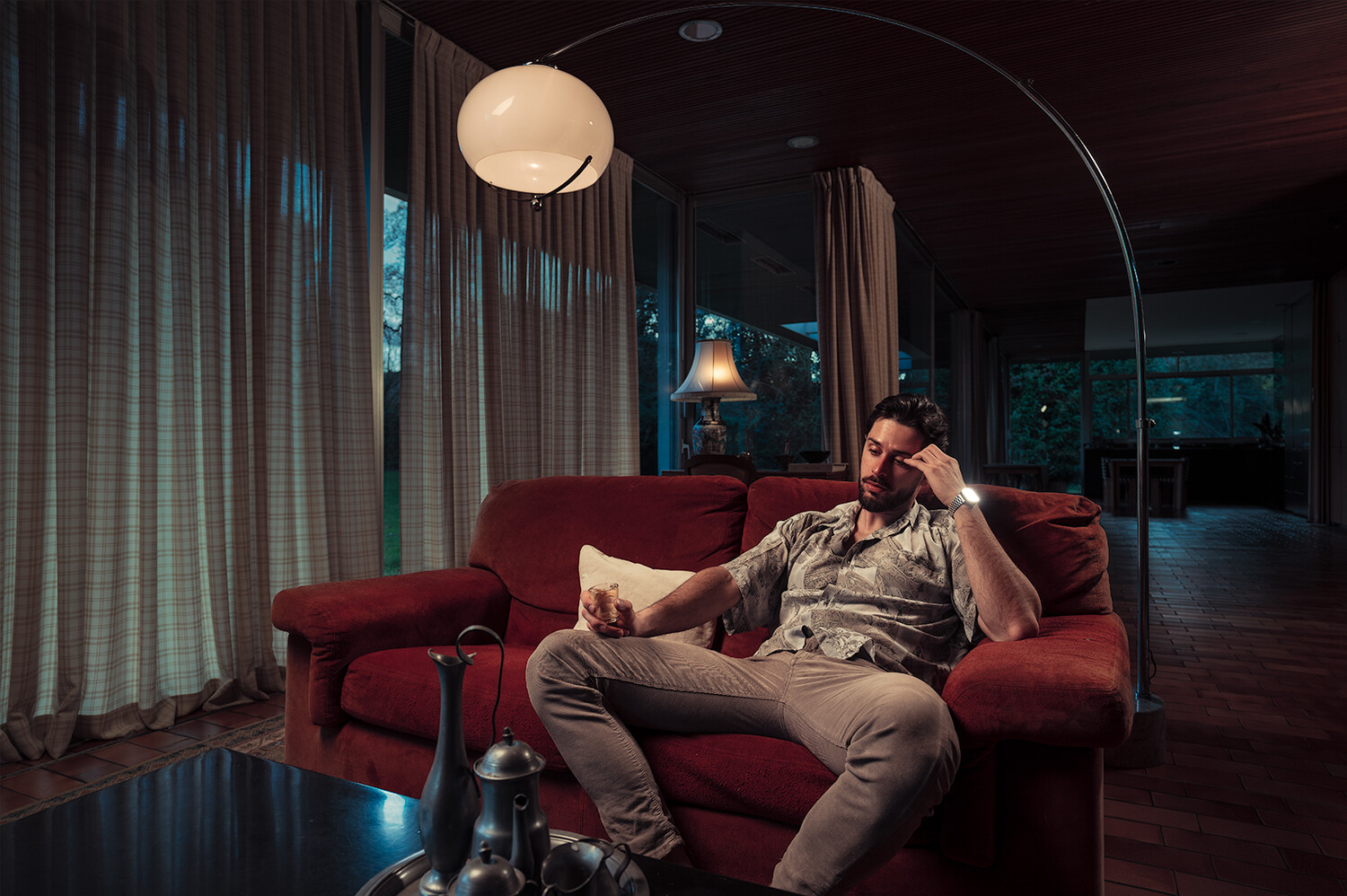Man relaxing on a burgundy sofa under an arc floor lamp in a modern interior at dusk