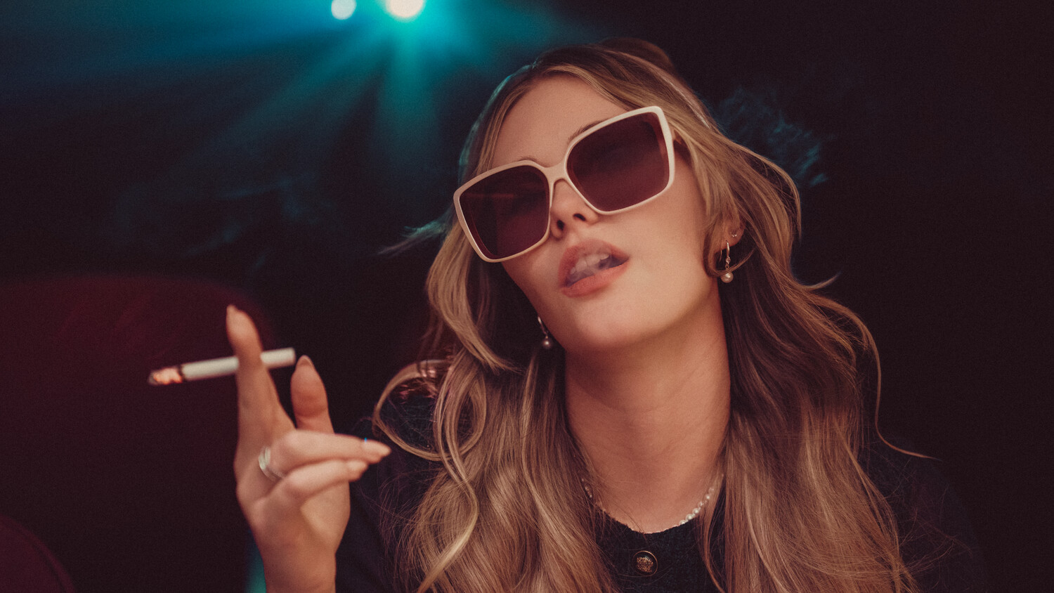 Woman wearing oversized white-framed sunglasses holds a cigarette while looking upward