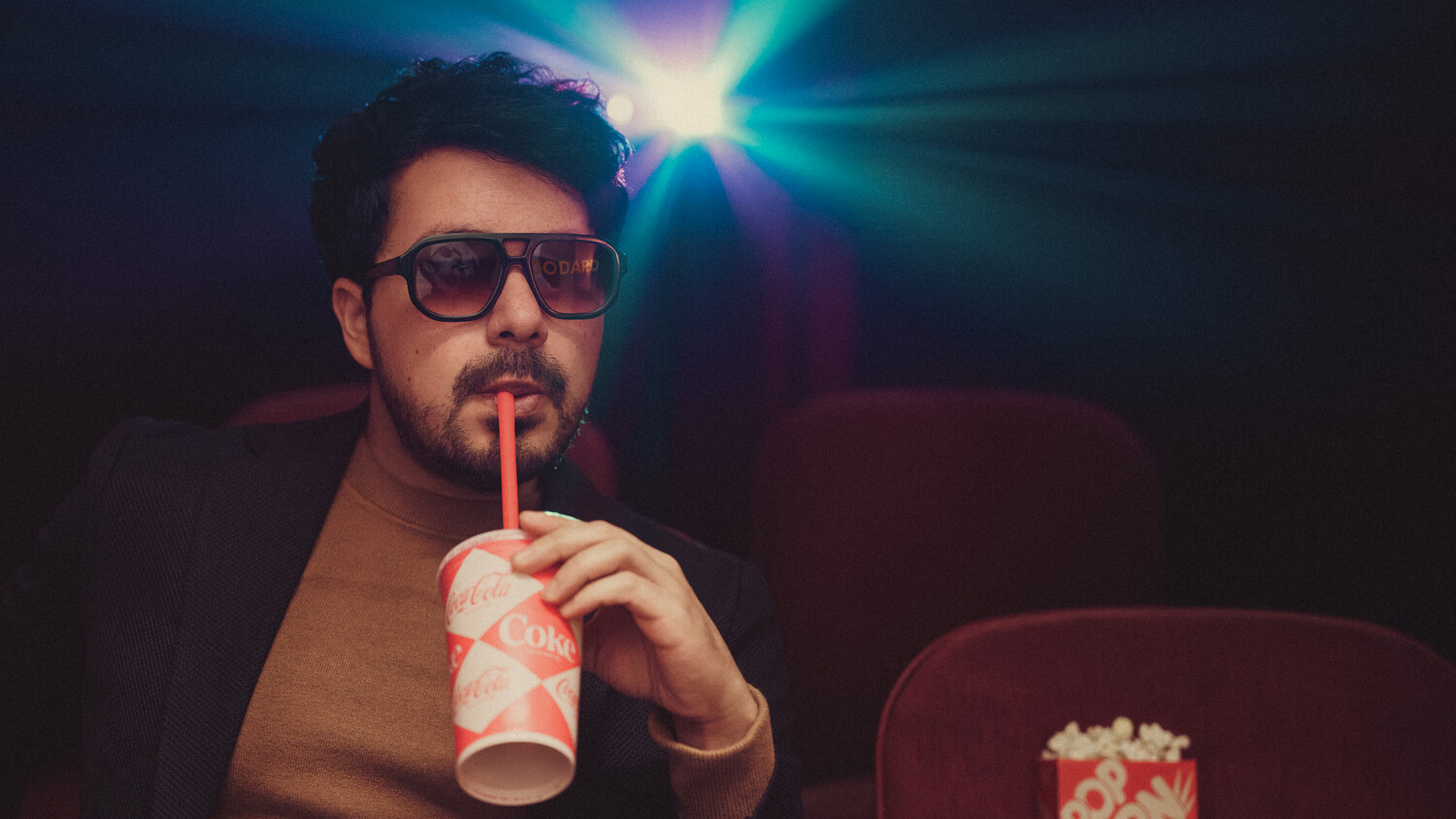 Man wearing sunglasses drinks from a straw in a darkened theater with colorful lens flare lighting