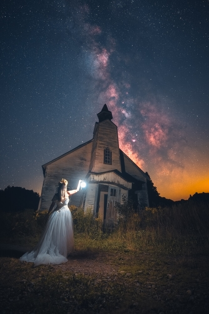 Woman in flowing white dress standing before abandoned church with Milky Way galaxy overhead