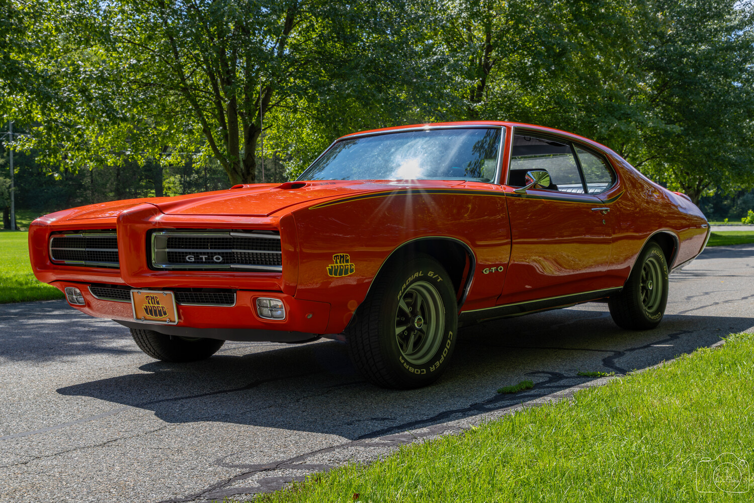 Bright red vintage muscle car parked on asphalt with lush green trees in background