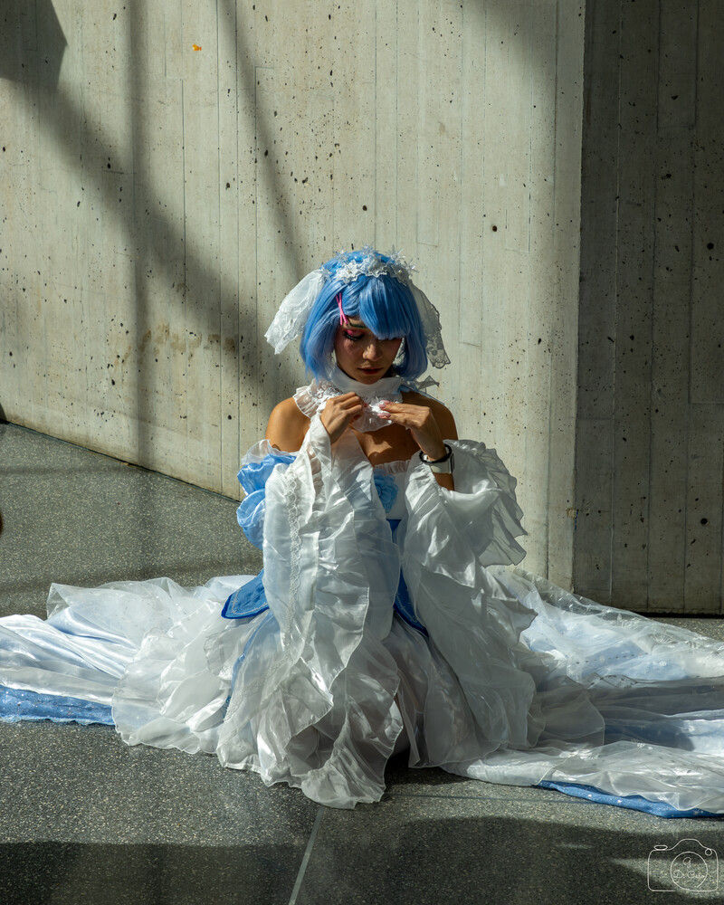 Cosplayer with blue hair and a white ruffled dress with blue accents, seated on pavement against a concrete wall and looking down