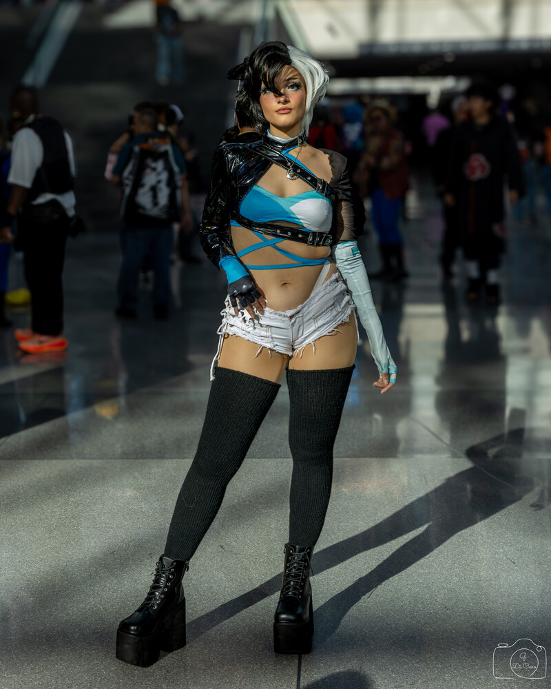 Full-length cosplayer in a black, blue, and white costume with platform boots and thigh-high stockings, standing on a glossy floor at a crowded convention.