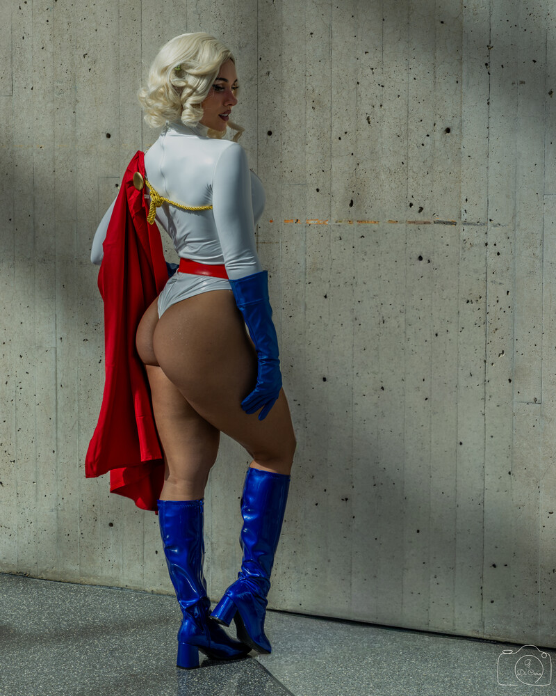 Cosplayer in a Power Girl costume with a flowing red cape and blue boots, photographed from behind against a concrete wall.
