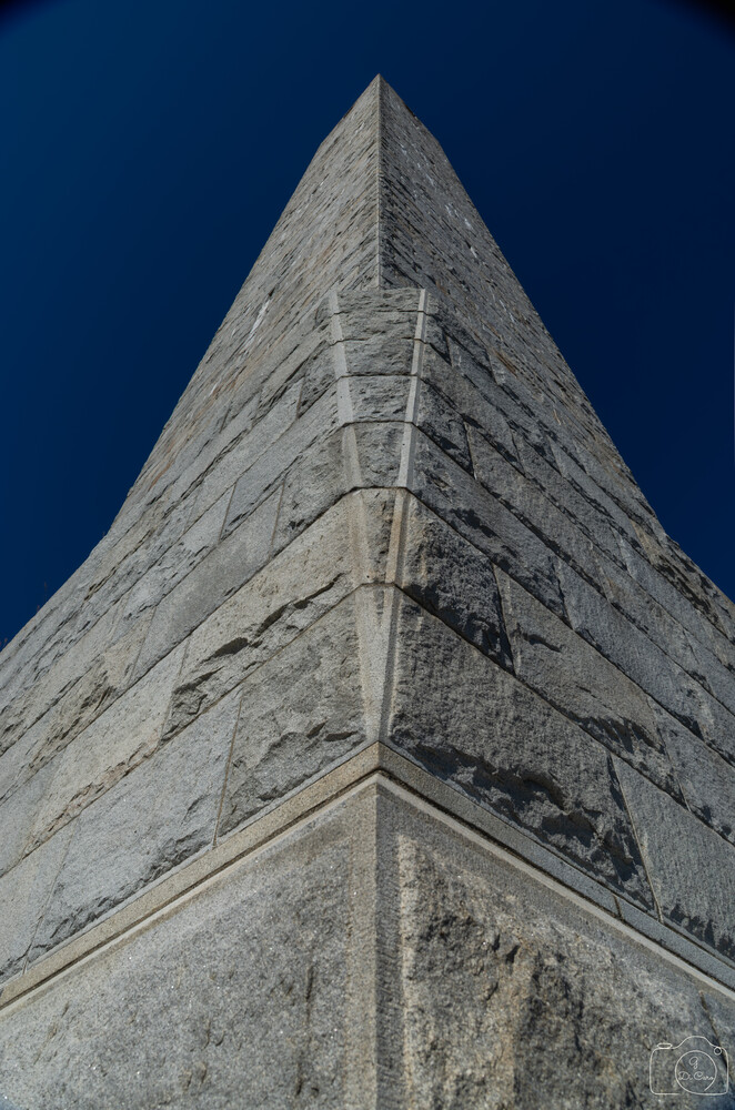 Low-angle view looking up at a tall stone obelisk with converging edges, set against a deep blue sky.