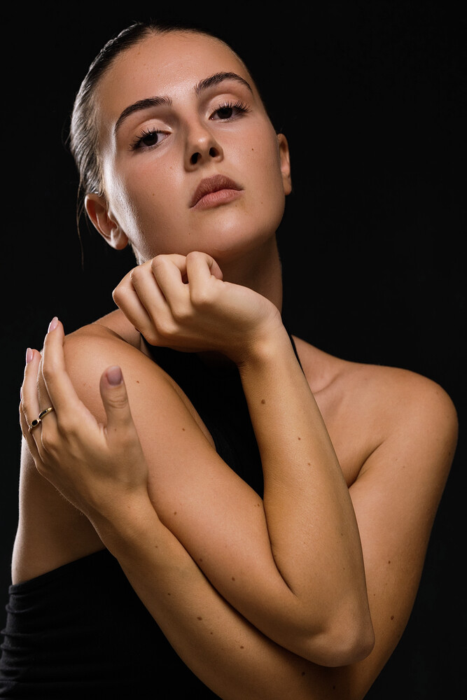 Woman with bare shoulders in studio portrait, hands near face, contemplative expression against black background