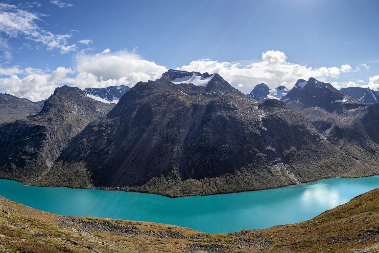 Turquoise glacial lake cradled by dark rocky mountains with snowy peaks under a partly cloudy blue sky.