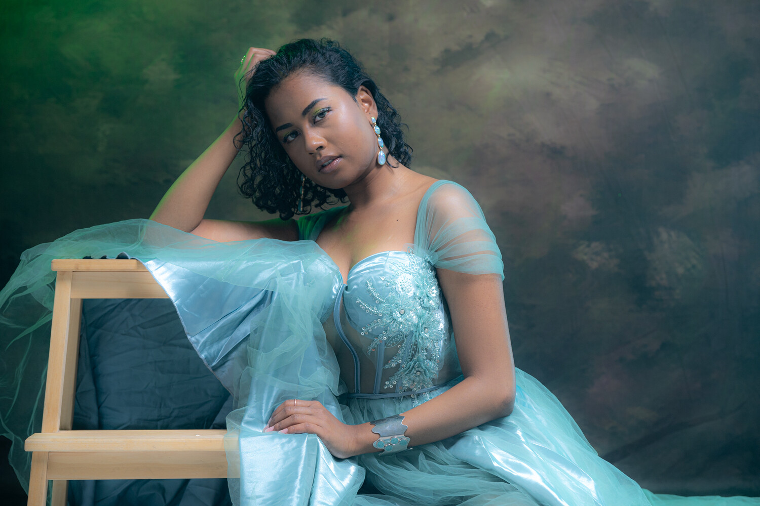 Woman in light blue tulle gown leaning against wooden chair against cloudy backdrop