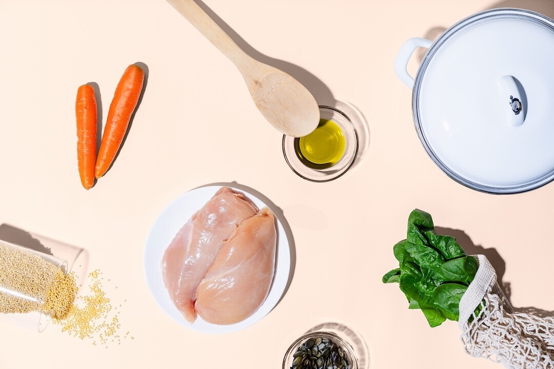 Flat lay food styling with raw chicken breast, carrots, spinach, olive oil, and cooking vessels on neutral background