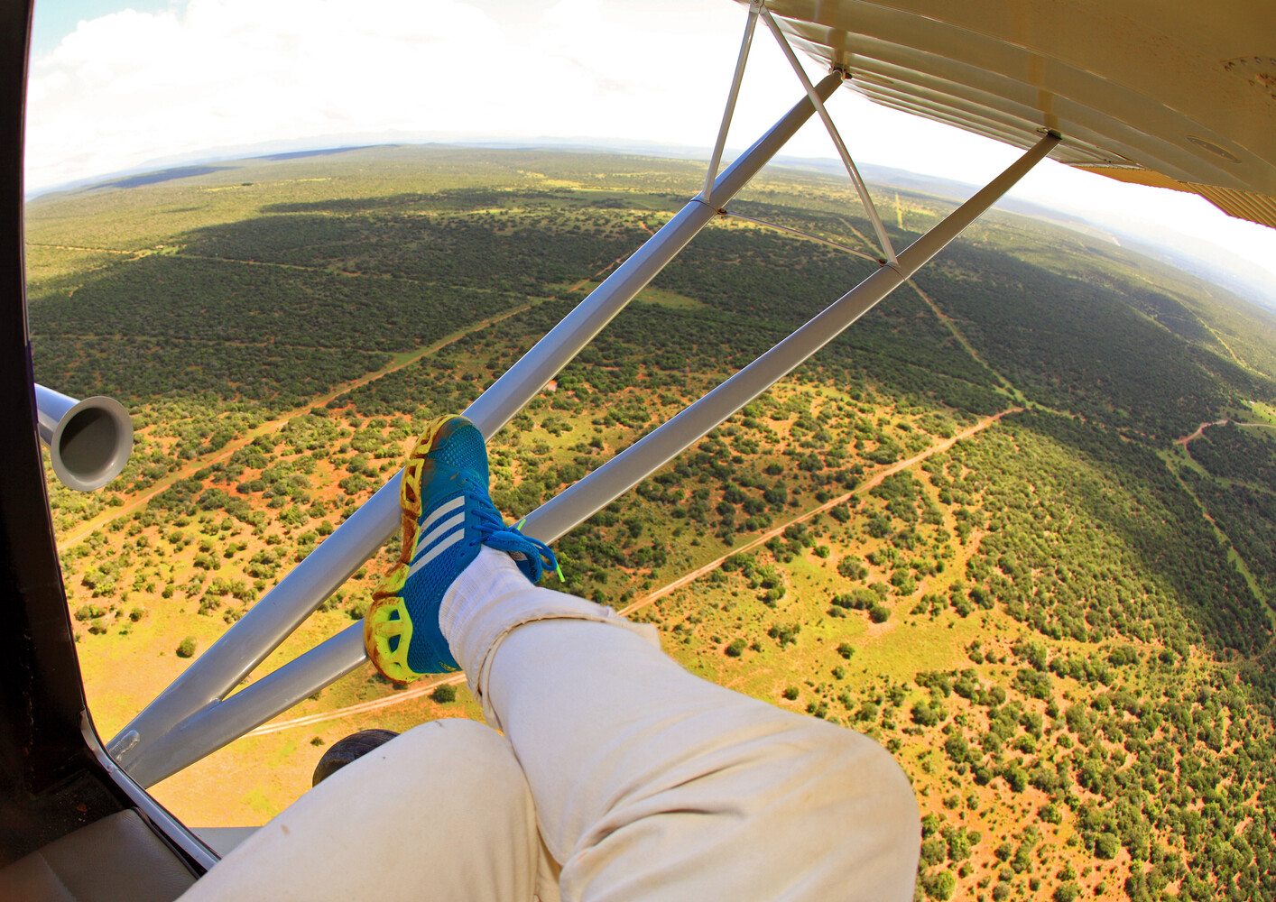 Pilot's leg and foot extending from open aircraft window with blue strut during aerial flight over forested landscape