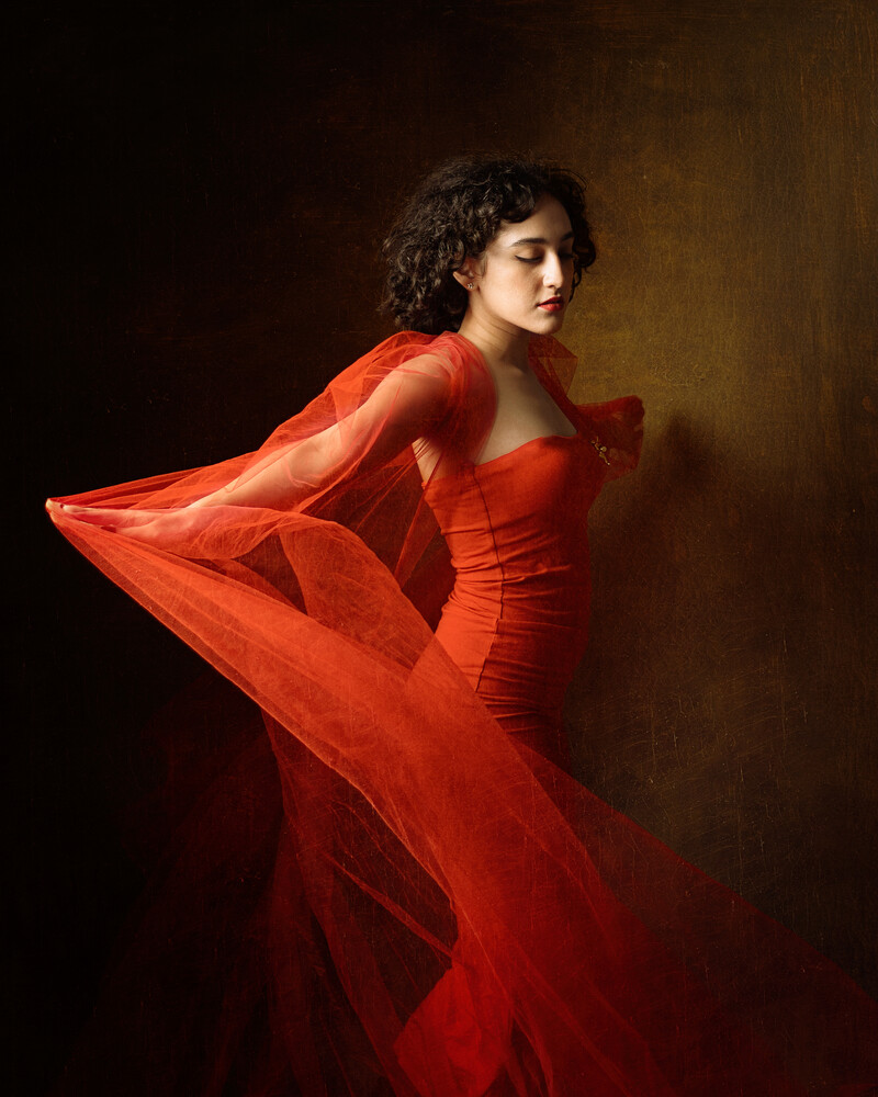 Woman in flowing red fabric against dark brown background, shot in profile