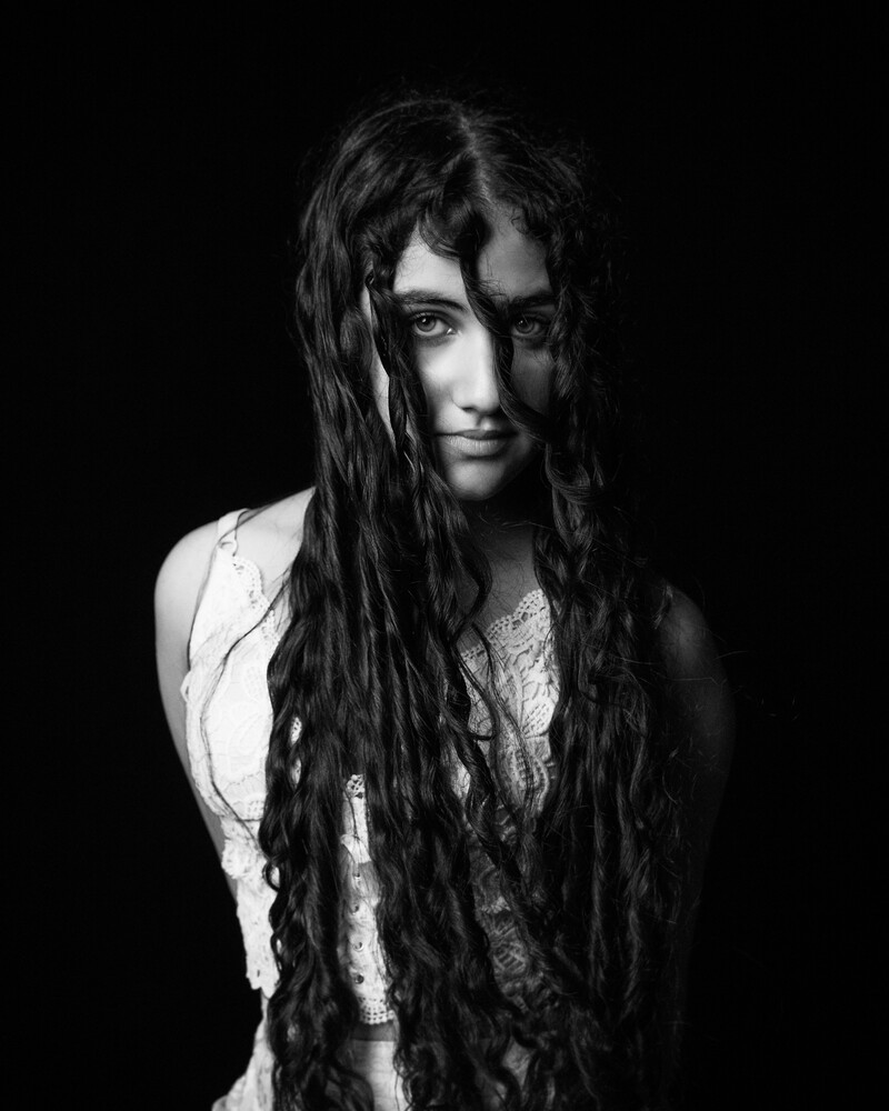 Black and white portrait of a woman with long wavy hair partly covering her face, against a black background.