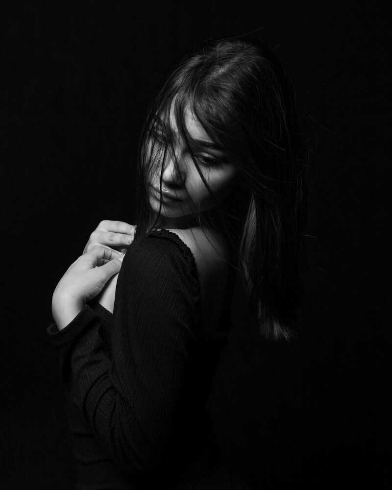 Black and white portrait of a woman with long hair falling across her face, one hand resting on her shoulder, against a solid black background.