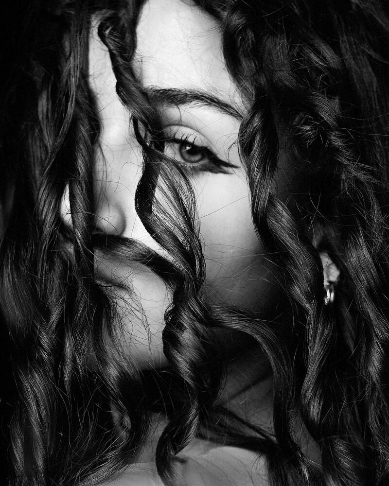 Black and white portrait of a woman with long, wavy hair falling across her face, with one dark eye and a raised hand visible.